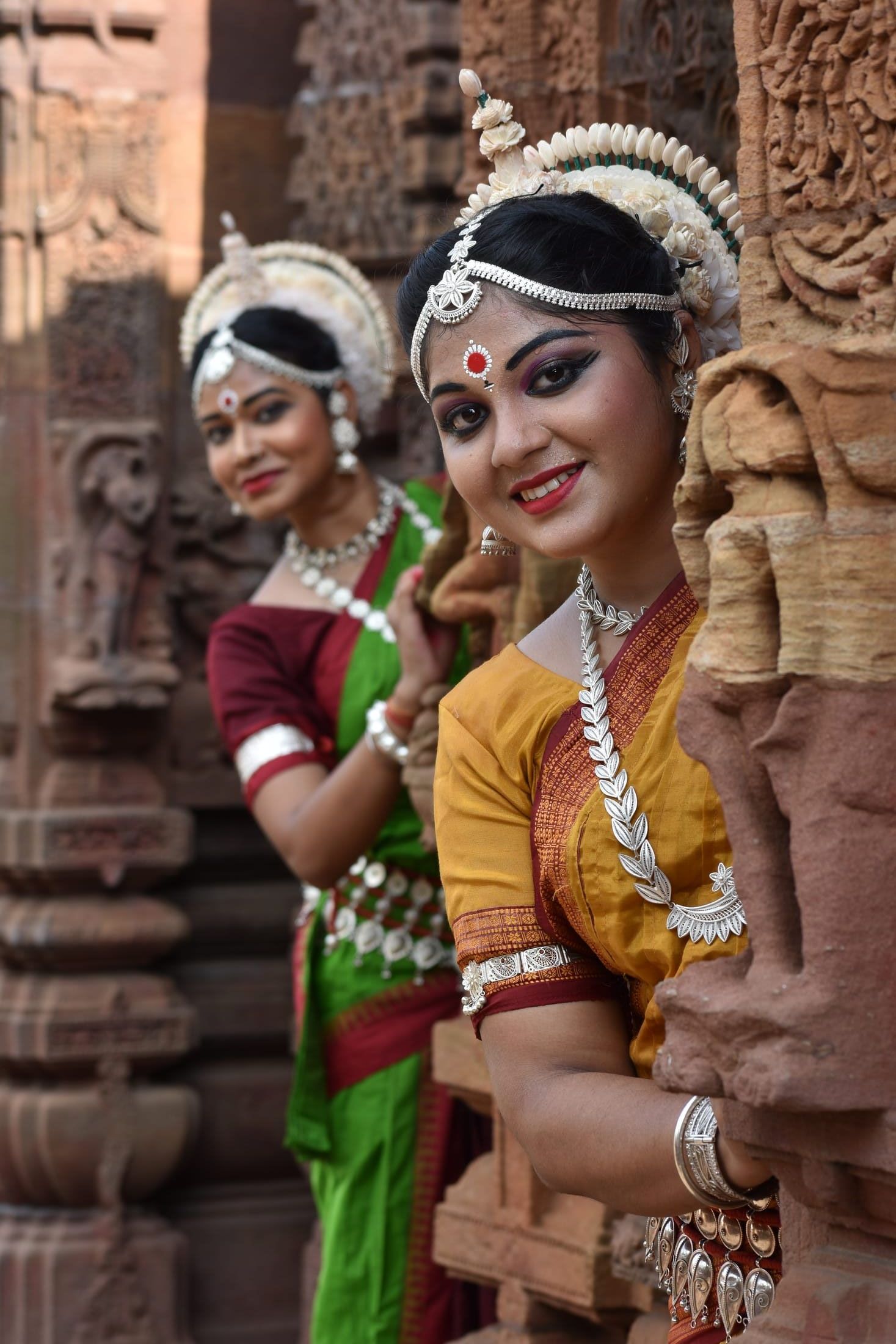 Two Odissi dancers reclining against carved temple stonework, smiling
