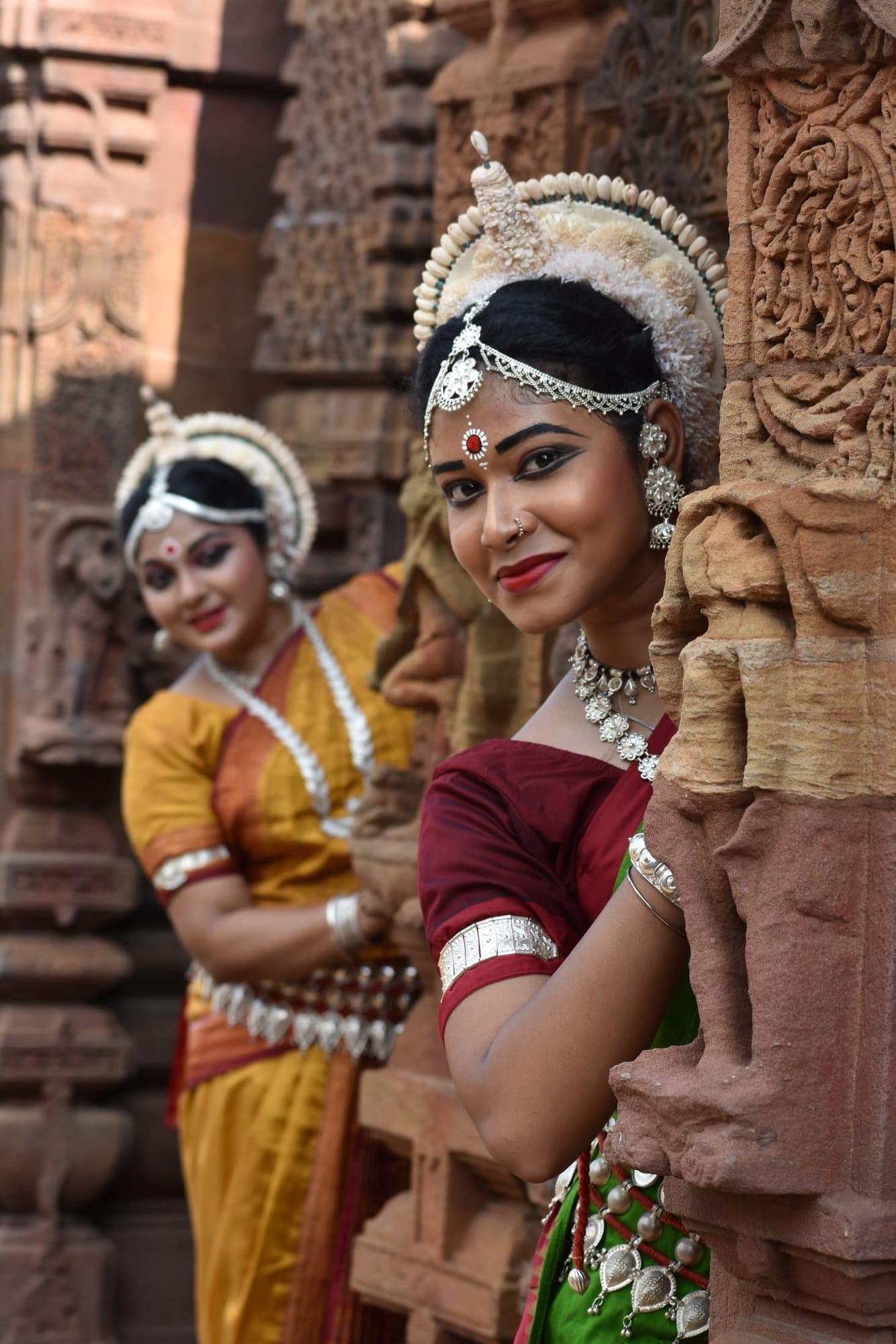 Two Odissi dancers smiling from behind carved temple pillars, candid moment