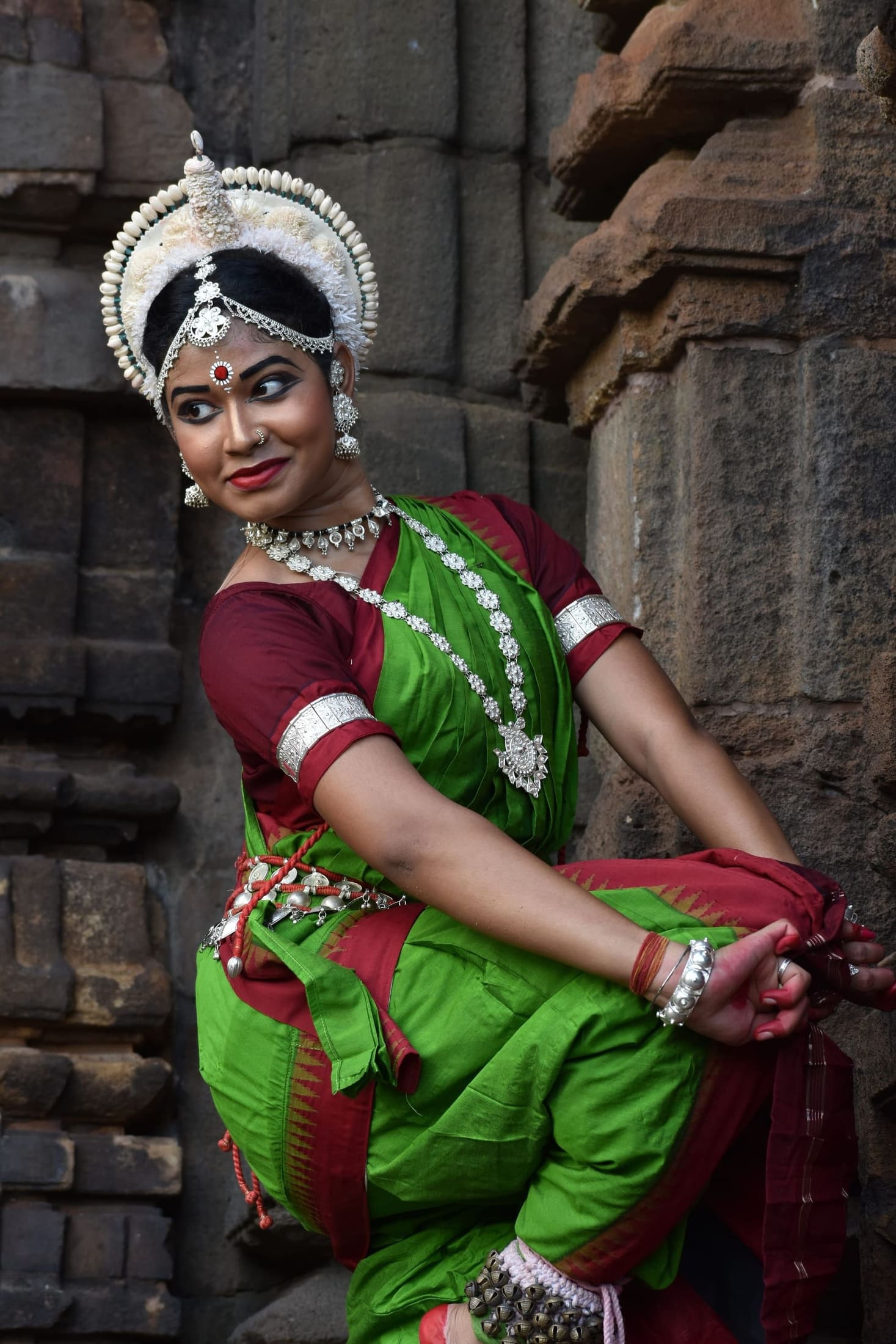 Odissi dancer in green costume with a playful glance against an ancient stone wall