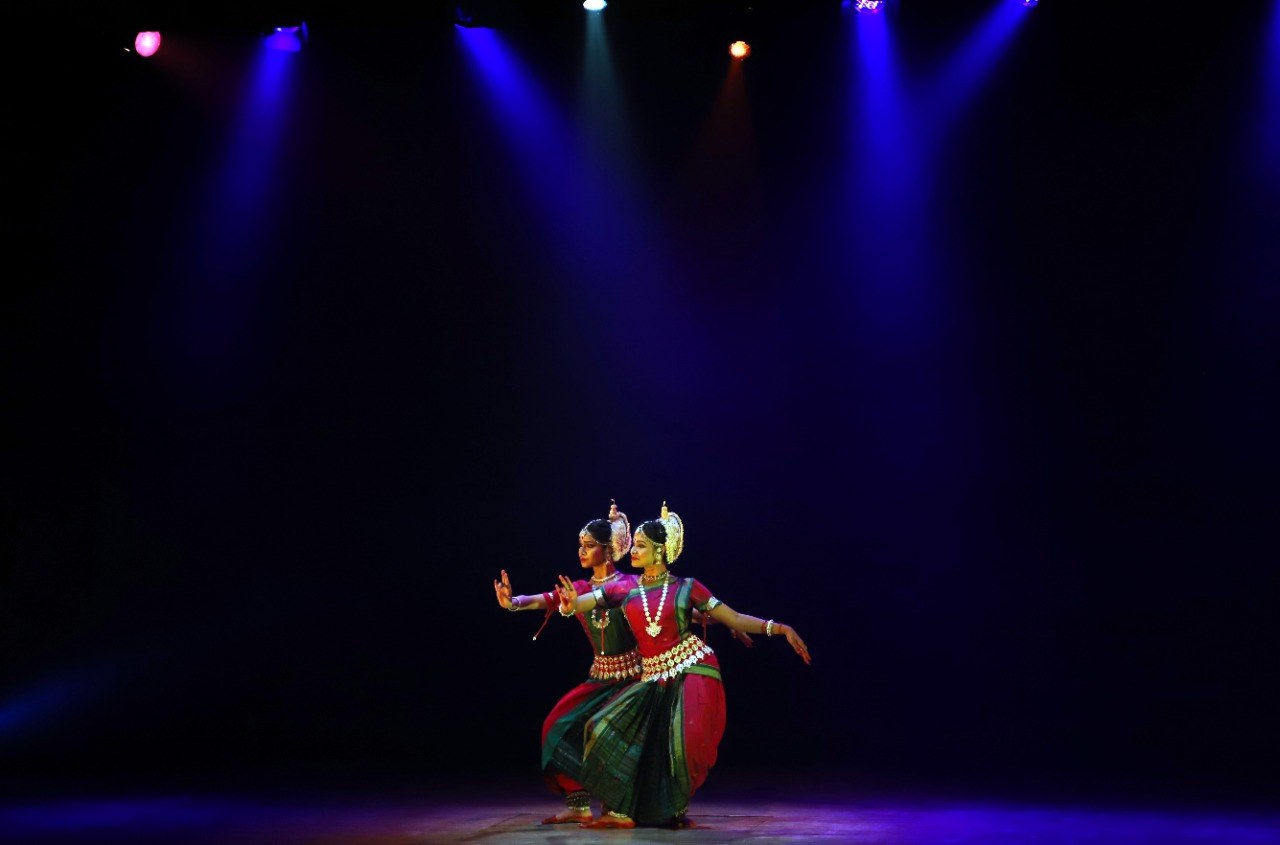 Two Odissi dancers in traditional pose under dramatic blue spotlight, wide empty stage