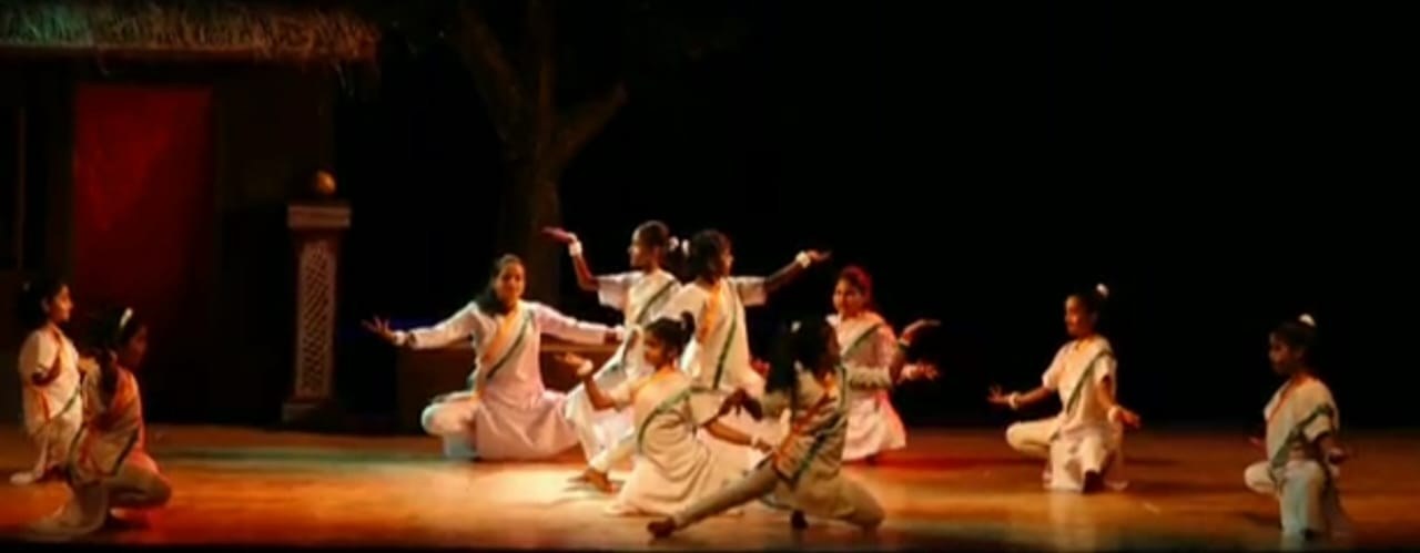Students in white patriotic costume performing on an outdoor stage with thatched backdrop
